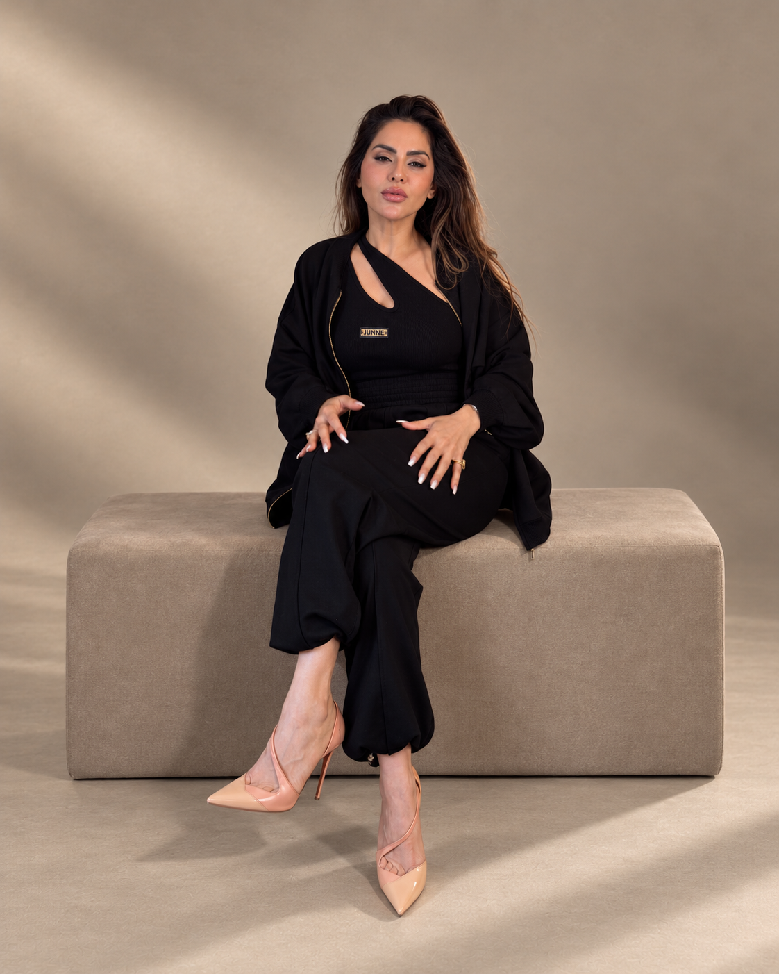 JUNNE Midnight Trio Co-ord Set — black Kylie Top, Kendall Jacket and Mila Joggers; seated front view on a taupe studio bench