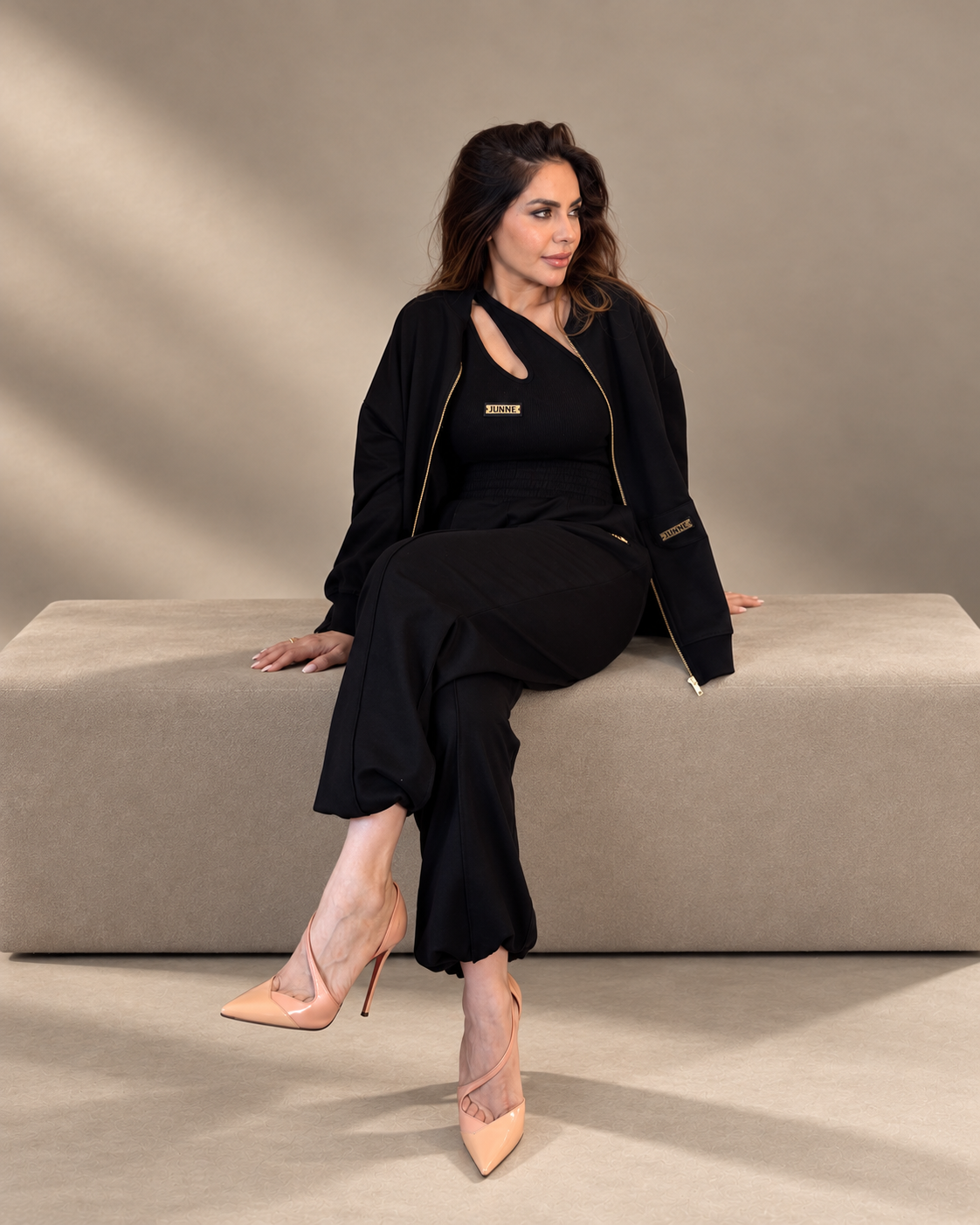JUNNE Midnight Trio Co-ord Set — black Kylie Top, Kendall Jacket and Mila Joggers; seated angled view on a taupe studio bench