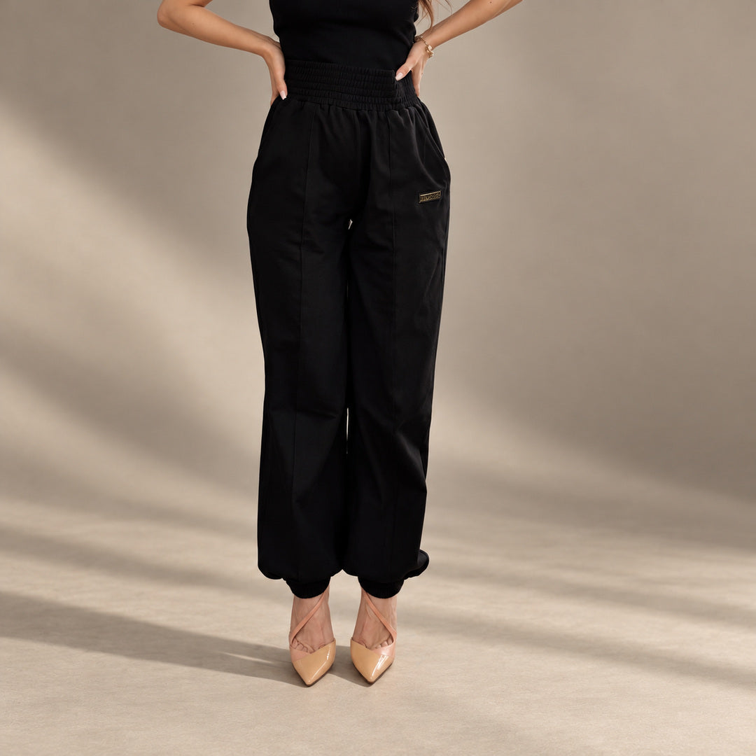 JUNNE Mila Black Pants — front view with wide smocked waistband and cuffed ankles on a minimal taupe studio background
