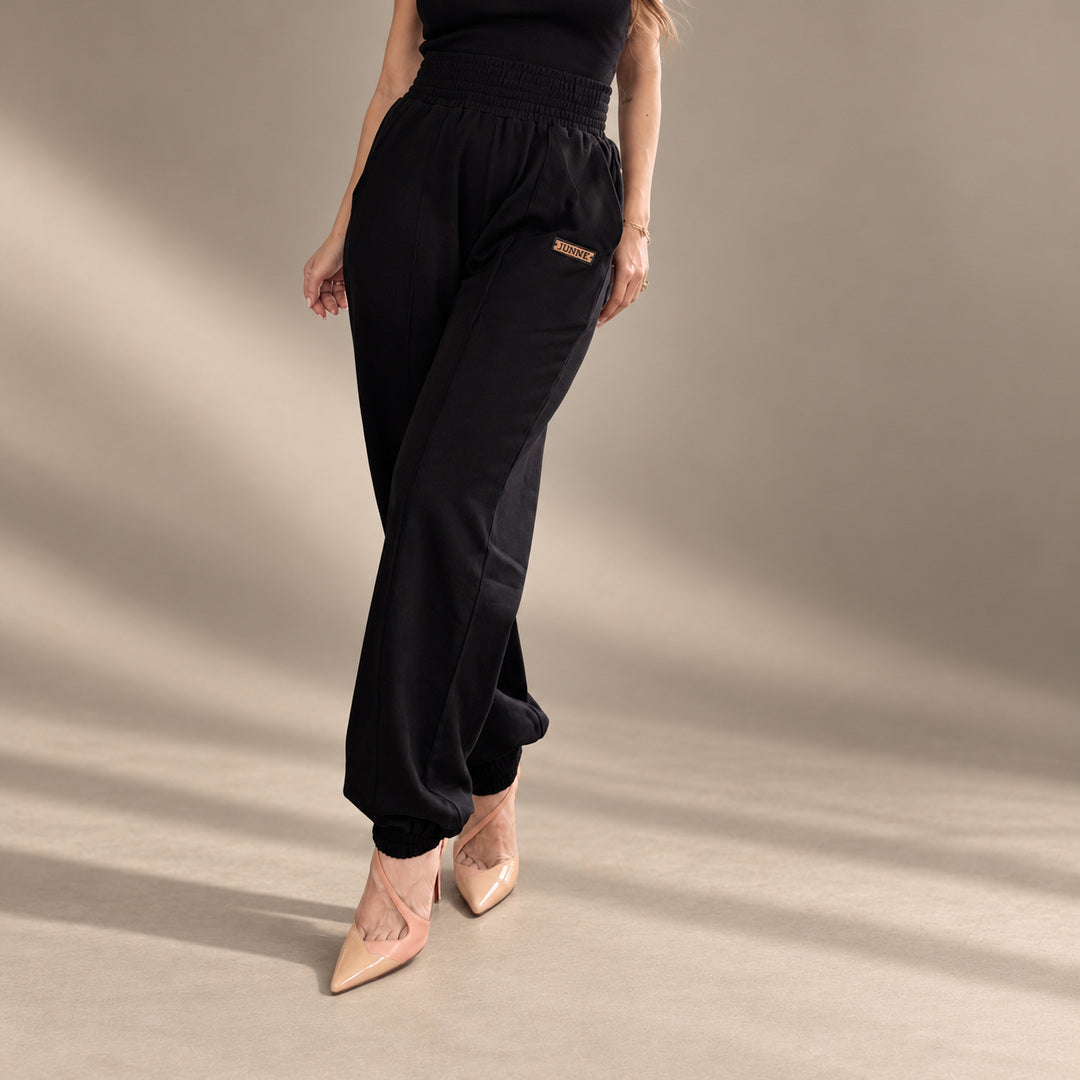 JUNNE Mila Black Pants — angled view showing relaxed drape and JUNNE branding on a minimal taupe studio background